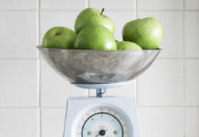 Een metalen kom gevuld met groene appels staat op een witte keukenweegschaal, met een betegelde witte muur op de achtergrond.
