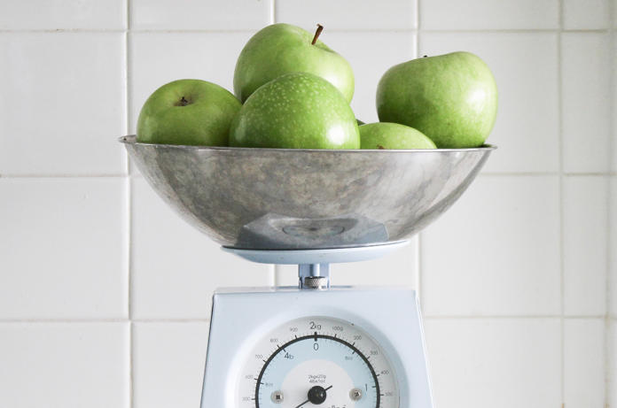 Een metalen kom gevuld met groene appels staat op een witte keukenweegschaal, met een betegelde witte muur op de achtergrond.