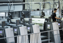 Een arbeider bedient een grote industriële drukpers in een moderne fabriek. Hij houdt toezicht op de bedrukte vellen terwijl deze door de verschillende stations van de machine gaan.