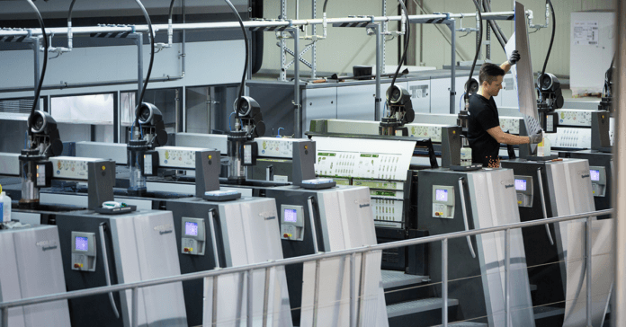 Een arbeider bedient een grote industriële drukpers in een moderne fabriek. Hij houdt toezicht op de bedrukte vellen terwijl deze door de verschillende stations van de machine gaan.