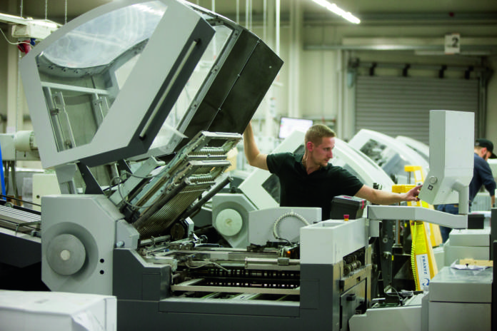 Een man bedient een grote industriële drukmachine in een fabriek en bedient de bedieningselementen terwijl hij omringd is door andere, vergelijkbare machines. De omgeving oogt schoon en goed verlicht.