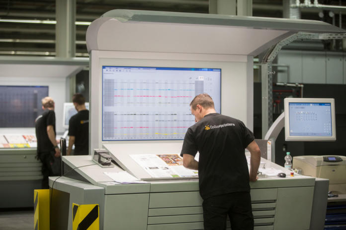 Een man staat bij een groot bedieningspaneel met schermen waarop grafieken en gegevens worden weergegeven in een drukkerij, terwijl op de achtergrond twee andere werknemers zichtbaar zijn.