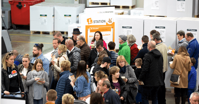 In een loodsachtige omgeving staat een grote groep mensen, bestaande uit zowel volwassenen als kinderen, in de rij. Velen houden papieren vast en kijken naar een station dat met een bord op de achtergrond wordt aangegeven.
