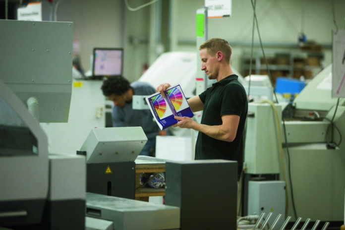 Een man in een zwart shirt bekijkt kleurrijk bedrukt papier in een moderne drukkerij vol grote machines; op de achtergrond is iemand anders aan het werk.