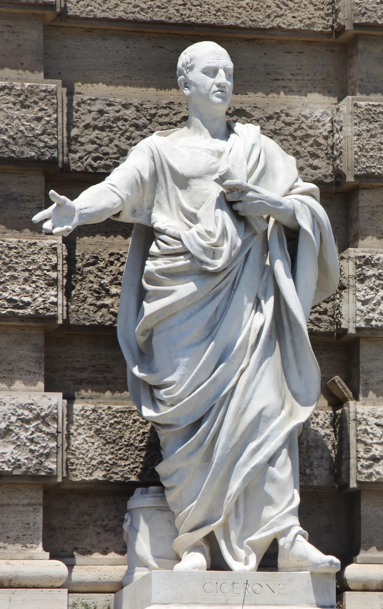 Standbeeld van een Romeinse redenaar in traditionele kledij, één arm gestrekt, met een rol in de andere hand. Het beeld staat voor een getextureerde stenen muur. Op een plaquette aan de voet staat CICERONE.