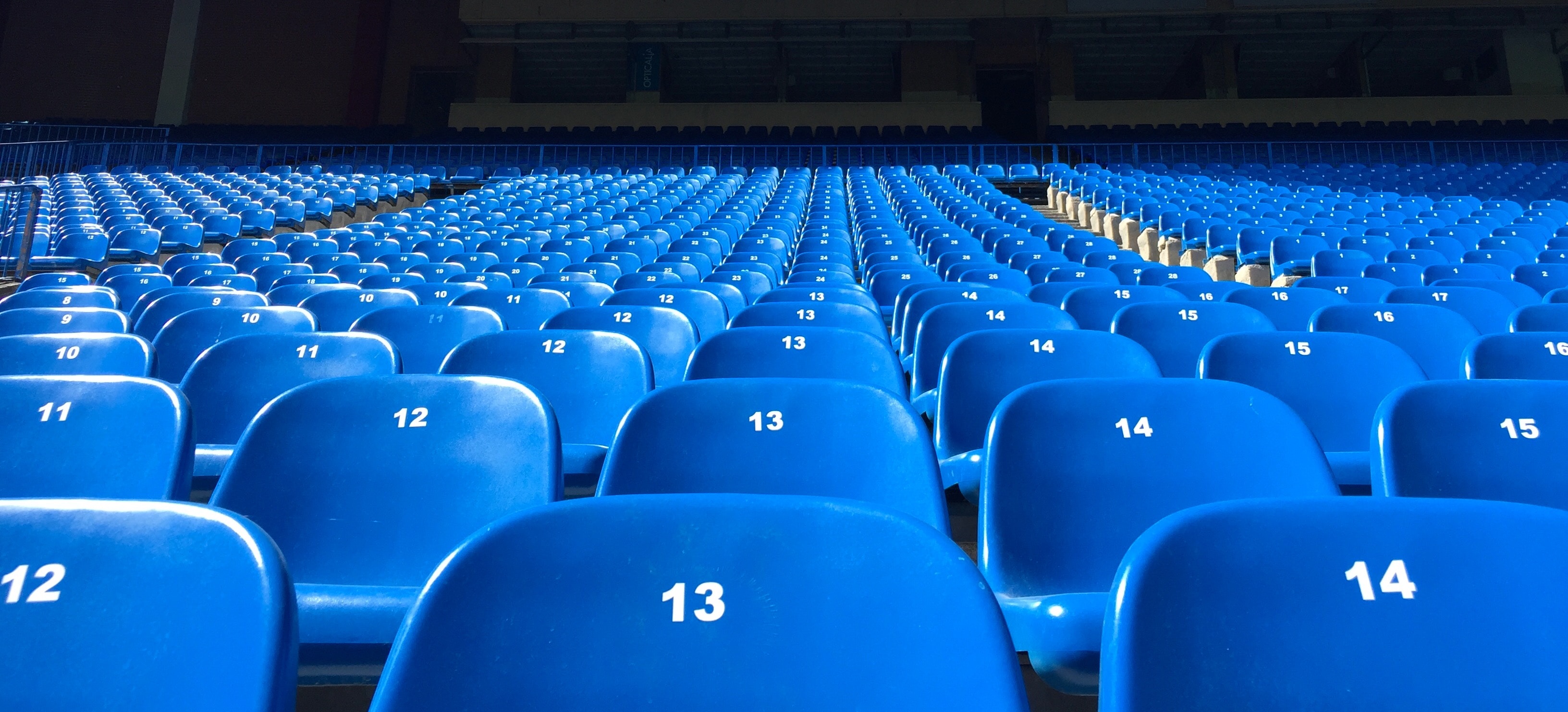 Een groot stadion met rijen lege blauwe stoelen, elk gemarkeerd met een nummer. De zichtbare nummers zijn 12, 13 en 14, terwijl het zonlicht over de stoelen speelt. Deze uitgestrekte tribune gonsde ooit van opwinding tijdens het EM 2021 Spielplan.