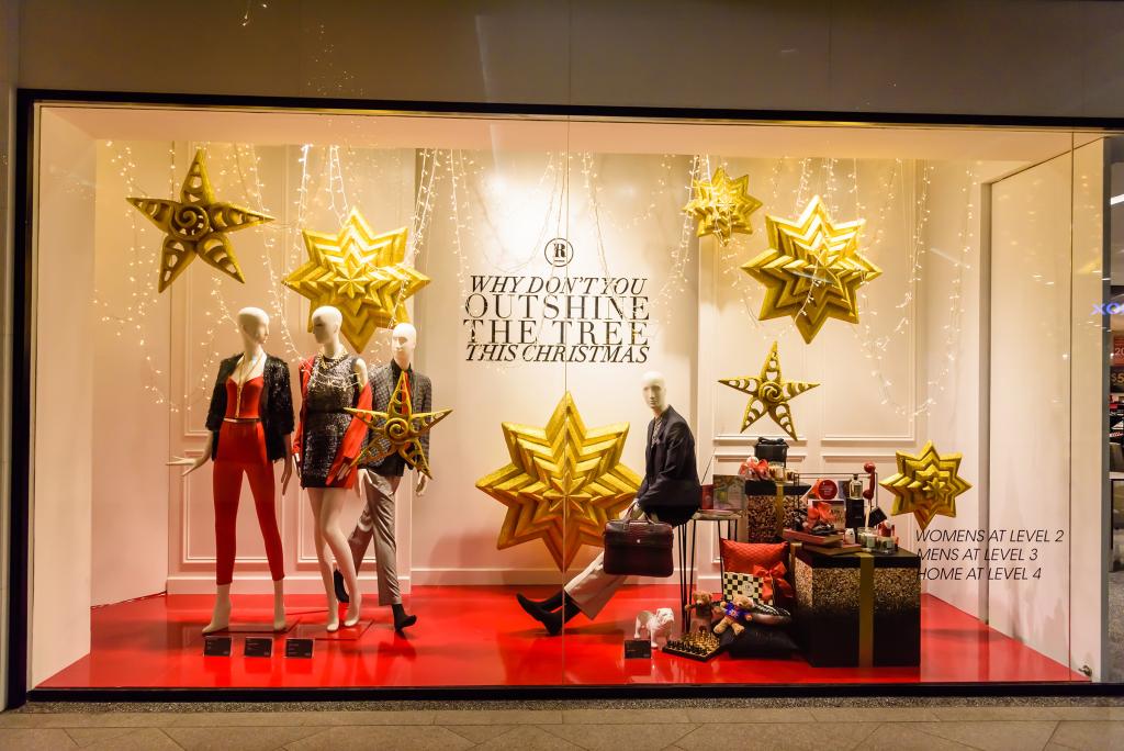 Etalage met vier mannequins in feestelijke kleding, omringd door grote gouden sterren. De tekst luidt: "Waarom laat je de boom deze kerst niet schitteren?". Stapels ingepakte cadeaus liggen op een rode vloer.
