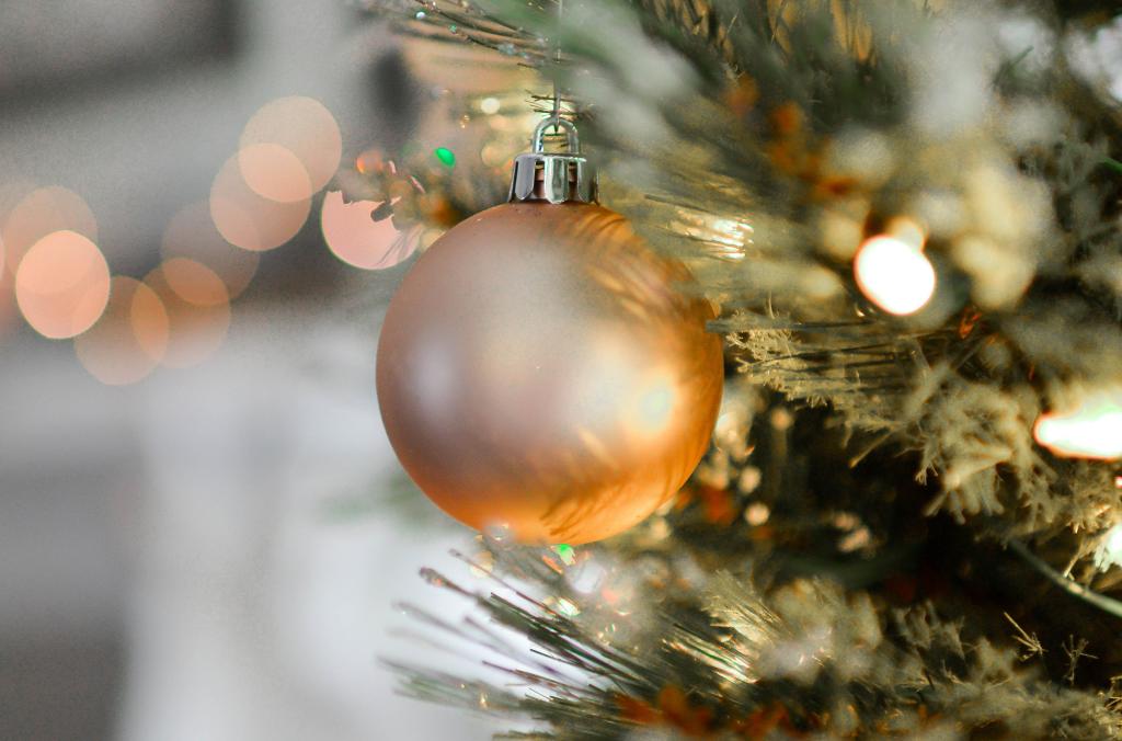 Close-up van een hangende matte gouden kerstbal in een boom met wazige, warme lichtjes op de achtergrond. De boom is versierd met groene naalden en zacht gloeiende lichtslingers, wat een feestelijke en gezellige sfeer creëert.