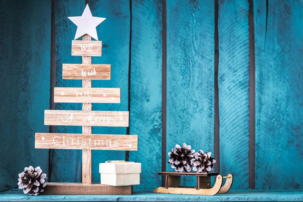Een houten kerstboom met een ster erop staat naast een klein geschenkdoosje. De boom heeft de tekst 'We Wish You A Merry Christmas'. Ernaast staat een miniatuurslee met dennenappels. De achtergrond bestaat uit blauwe houten planken.