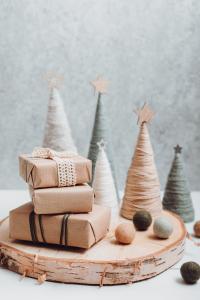 Een feestelijk tafereel met kleine ingepakte cadeautjes op een rustieke houten voet, omringd door met touw omhulde kegelvormige bomen met sterren erop. Er liggen verspreid viltballetjes en de achtergrond heeft een getextureerd, grijs oppervlak.