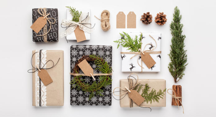 Een verzameling ingepakte cadeaus op een witte ondergrond. De pakjes zijn versierd met groen, touw, dennenappels en kaneelstokjes. Aan elk cadeau zitten labels bevestigd. De verpakkingen zijn versierd met patronen zoals sneeuwvlokken en rendieren.
