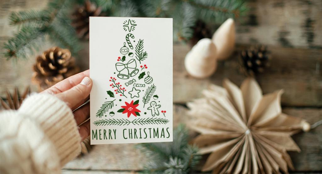 Een hand houdt een kaart vast met een decoratieve kerstboom, gemaakt van kerstsymbolen zoals ornamenten, zuurstokken en peperkoek. Daaronder staat 'Vrolijk Kerstfeest'. De achtergrond is versierd met feestelijk groen en dennenappels.