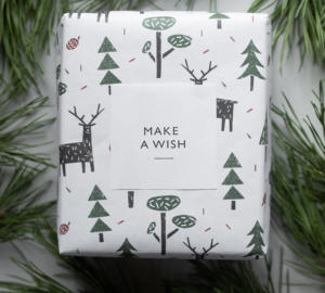Een cadeau verpakt in wit papier met groene bomen en zwarte herten. De verpakking heeft een label met de tekst MAKE A WISH. Het cadeau is omringd door dennentakken.