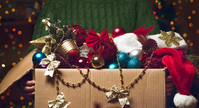Een persoon in een groene trui houdt een open kartonnen doos vast, gevuld met kerstversiering, waaronder kerstballen, een kerstmuts, kerstversiering en kleine strikjes. De achtergrond is versierd met feestelijke, wazige lichtjes.