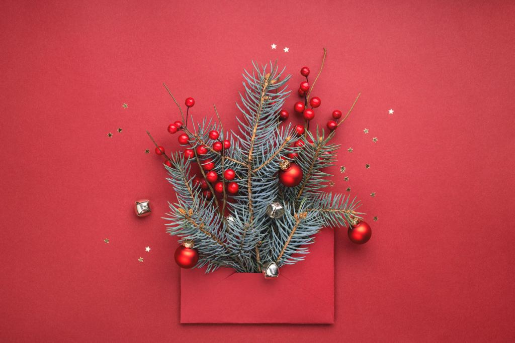 Feestelijk arrangement op een rode achtergrond met een rode envelop met groenblijvende takken, rode bessen en rode ornamenten. Zilveren belletjes en kleine gouden sterretjes zijn verspreid.
