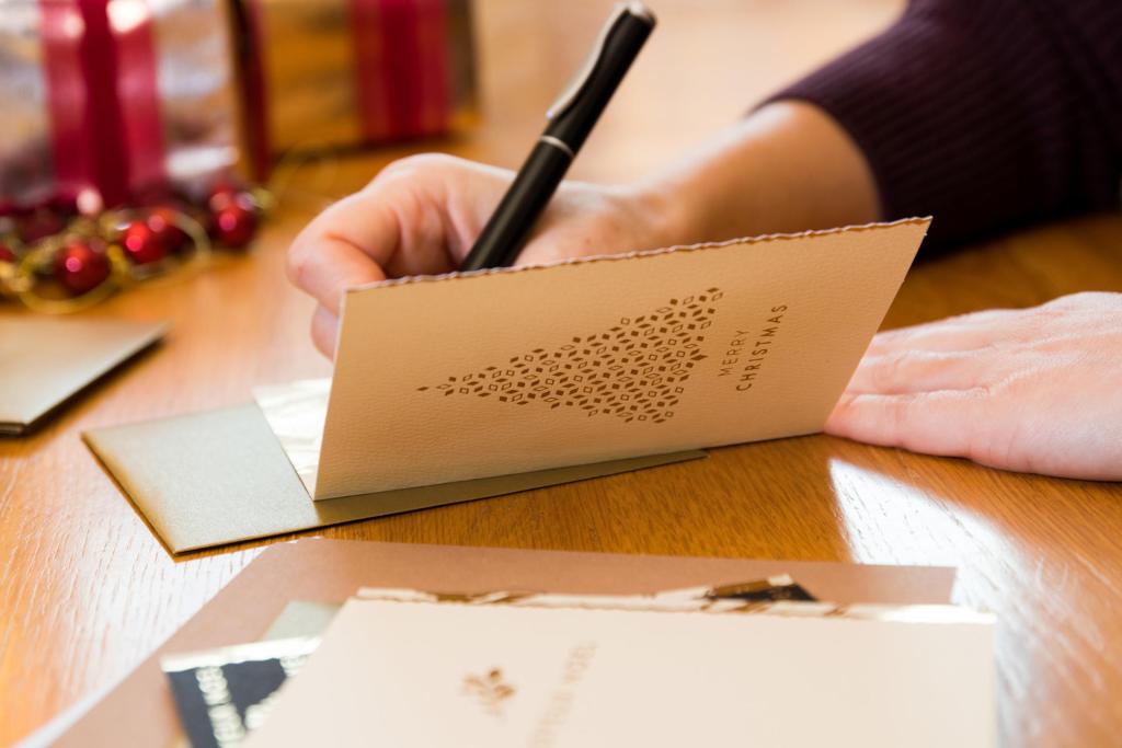 Iemand schrijft met een zwarte pen op een kerstkaart. De kaart is versierd met een gestileerde kerstboom en de tekst Merry Christmas. Er liggen enveloppen en nog een kaart op de houten tafel.