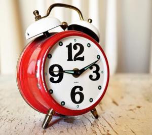 Een vintage rode wekker met twee bellen bovenop, die de tijd aangeven als 10:10. De klok heeft een witte wijzerplaat met grote zwarte cijfers en staat op een lichtgekleurd oppervlak met textuur. Op de achtergrond zijn vage witte gordijnen te zien.