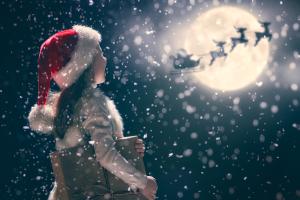 Een kind met een kerstmuts houdt een cadeautje vast en kijkt naar de nachtelijke hemel vol sneeuwvlokken. De volle maan is zichtbaar en het silhouet van een door rendieren getrokken slee is daartegen afgetekend, wat een magisch, feestelijk tafereel oproept.