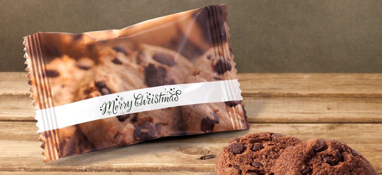 Een ingepakt koekje met chocoladeschilfers, met een vrolijk kerstlabel, op een houten ondergrond. Twee chocoladekoekjes liggen naast de verpakking. De achtergrond is zacht, gedempt groen.