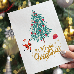 Een hand houdt een kerstkaart vast met een versierde boom, een klein kerstmanfiguurtje en een rood geschenkdoosje. De woorden Merry Christmas staan er in gouden letters op. Op de achtergrond is een wazige kerstboom te zien.